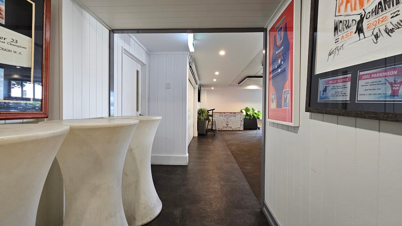 On the left, there are tall, white pedestal tables and a white panelled wall with framed pictures. In the middle, there is a hallway with dark flooring leading to a well-lit area with plants and a white table. In the background, there is a white wall with a door and more plants. On the right, there is a white panelled wall with framed posters and a bulletin board. The ceiling is white with recessed lighting.