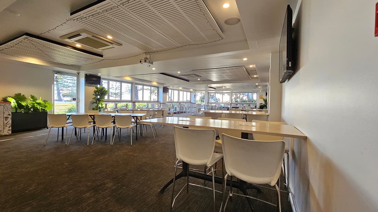 On the left, there are several tables with white chairs and potted plants near the windows. In the middle, there are more tables and chairs arranged in rows, with large windows in the background letting in natural light. On the right, there is a white wall with a mounted TV and a long table with white chairs. The floor is covered in dark carpet, and the ceiling has white panels with recessed lighting and air conditioning units.