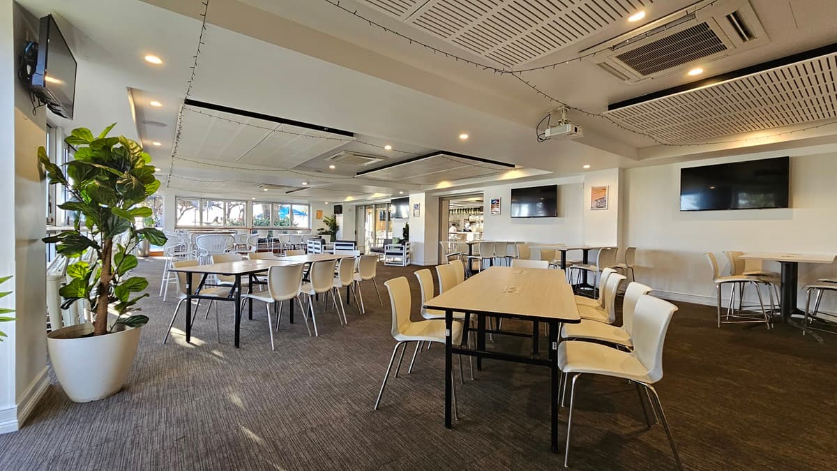 On the left, there are several tables with white chairs and a large potted plant near the windows. In the middle, there are more tables and chairs arranged in rows, with large windows in the background letting in natural light. On the right, there are two wall-mounted TVs above a long table with white chairs. The floor is covered in dark carpet, and the ceiling has white panels with recessed lighting and air conditioning units.