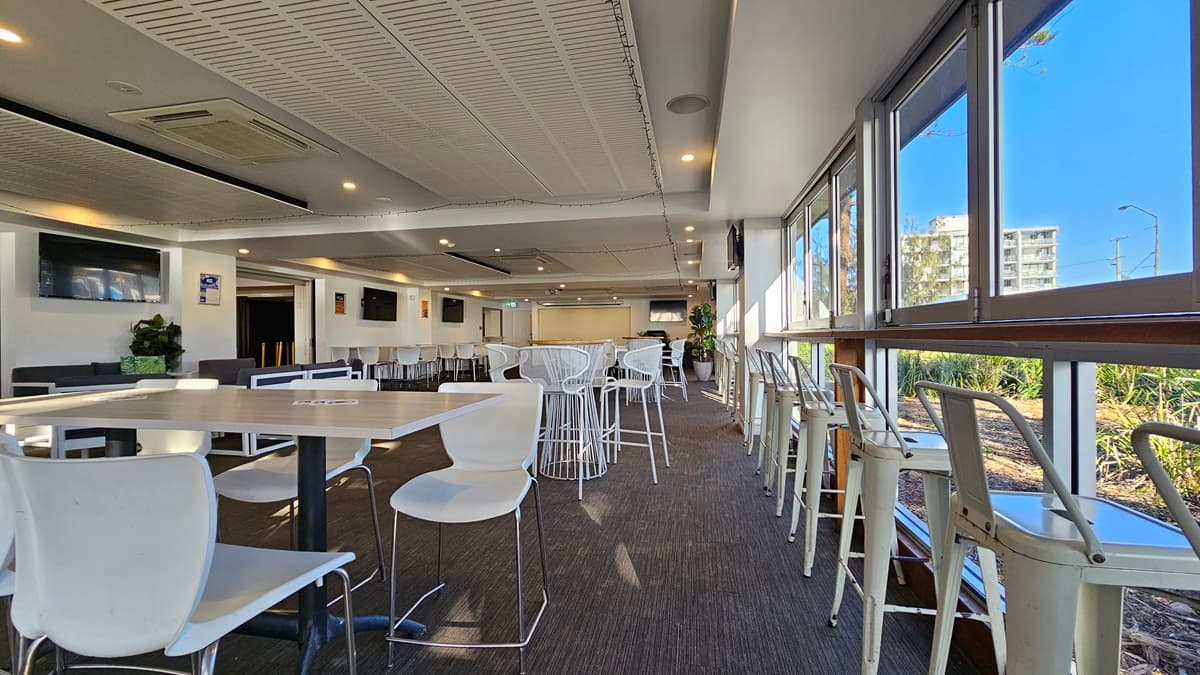 On the left, there are white tables with white chairs and a wall-mounted TV above a potted plant. In the middle, there are more white tables and chairs, with a long counter and bar stools in the background. On the right, there are large windows with high stools and tables along the window line, offering a view of greenery outside. The floor is covered in dark carpet, and the ceiling has white panels with recessed lighting and air conditioning units.