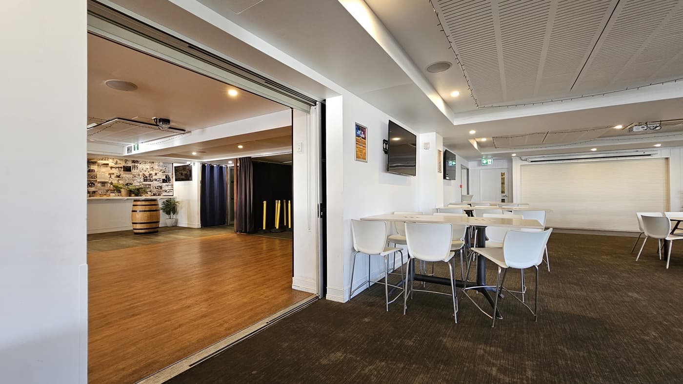 On the left, there is an open area with wooden flooring, a large barrel, and a wall covered in photos. In the middle, there is a partition wall separating the wooden floor area from the carpeted area. In the foreground, there are white tables and chairs on dark carpet. On the right, there is a wall-mounted TV above a white table and chairs, and a large roller door in the background. The ceiling has white panels with recessed lighting and air conditioning vents.