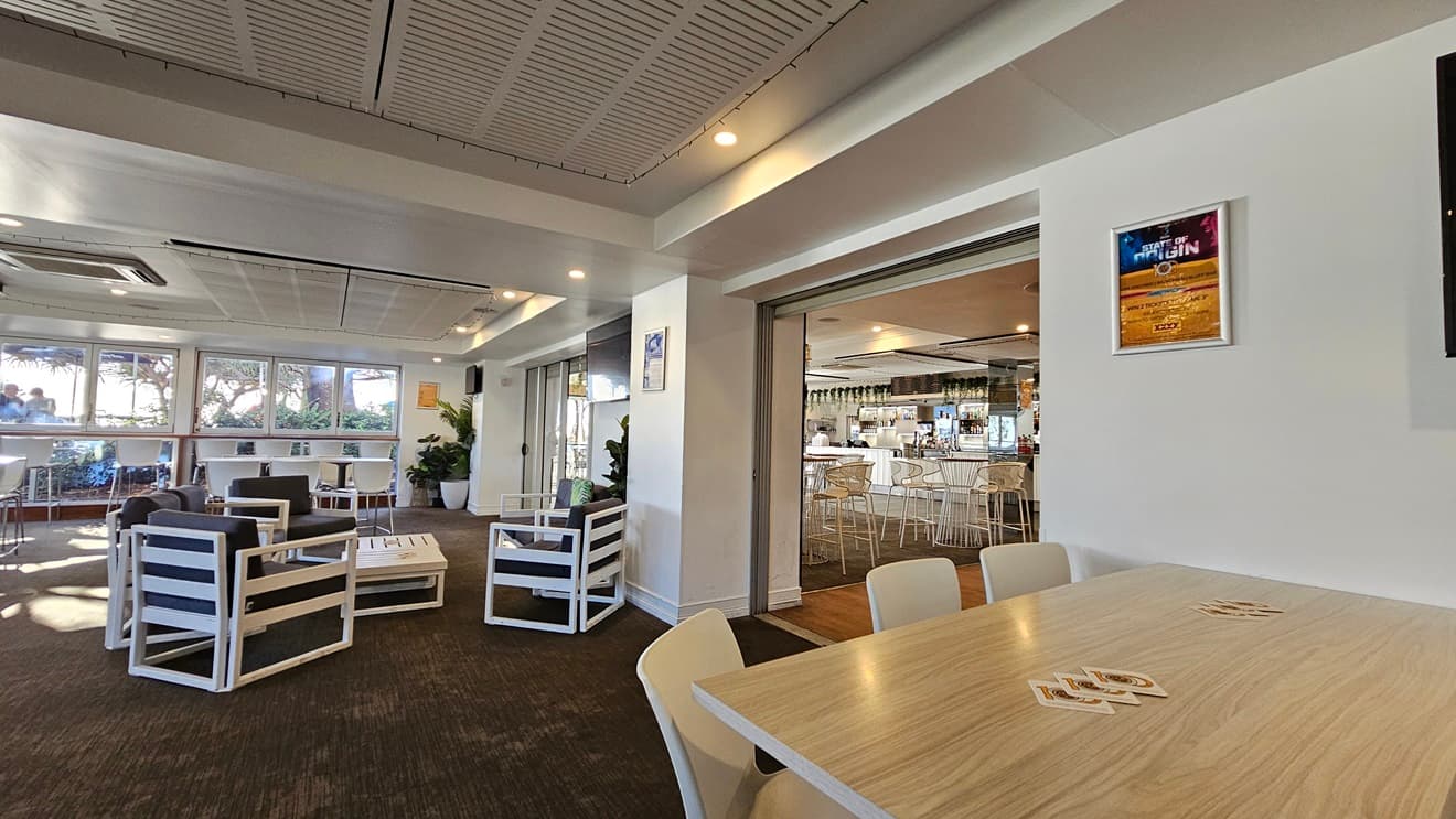On the left, there are white-framed lounge chairs with dark cushions around low tables, and large windows with a view of trees. In the middle, there is a partition wall with a doorway leading to a bar area in the background, featuring high stools and a counter. On the right, there is a wooden table with white chairs and a framed poster on the wall. The floor is covered in dark carpet, and the ceiling has white panels with recessed lighting and air conditioning vents.
