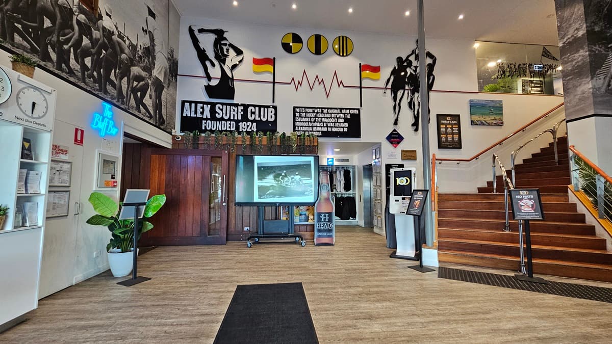 On the left, there is a wooden door with a "Bar" sign above it, a clock, and a potted plant. In the middle, a large screen on a stand displays an image, with surf club memorabilia and wall art above it. On the right, a staircase with wooden steps and a metal handrail leads to an upper level. The background includes more surf-themed decorations and a glass railing. The floor is wooden with a black mat in the centre, and the ceiling has recessed lighting.