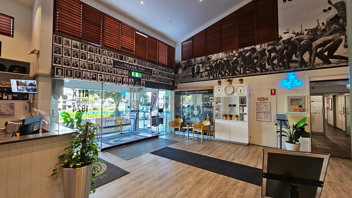 On the left, there is a reception desk with a plant and a computer monitor displaying security footage. In the middle, there are two yellow chairs with cushions and a small round table between them, in front of a glass display case filled with trophies and historical photos. On the right, there is a wooden door with a "Bar" sign above it, a potted plant, and a hallway leading to other rooms. The floor is a mix of wooden and carpeted areas, and the ceiling has recessed lighting and wooden blinds.