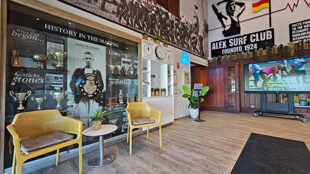 On the left, there are two yellow chairs with cushions and a small round table between them, in front of a glass display case filled with trophies and historical photos. In the middle, there is a wall with clocks and a potted plant. On the right, a large screen displays a beach scene, next to a wooden door with a "Bar" sign above it. The background features surf club memorabilia and wall art. The floor is wooden with a black mat in the centre, and the ceiling has recessed lighting.