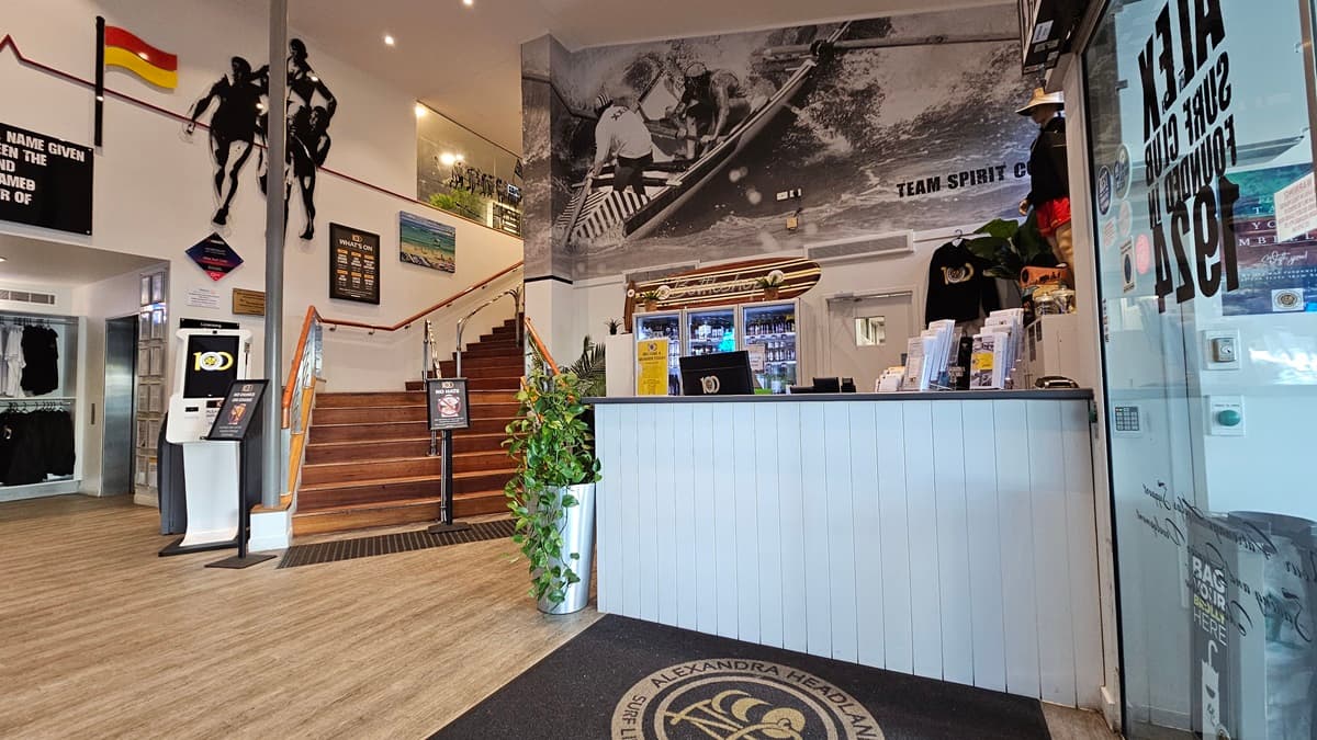 On the left, there is a staircase with wooden steps and a metal handrail, leading to an upper level. In the middle, there is a wall with surf-themed artwork and a large black-and-white mural of surfers above a reception counter. On the right, the reception counter has a plant, merchandise, and a display case with drinks. The floor is wooden with a black mat in front of the counter, and the ceiling has recessed lighting and surf club flags.