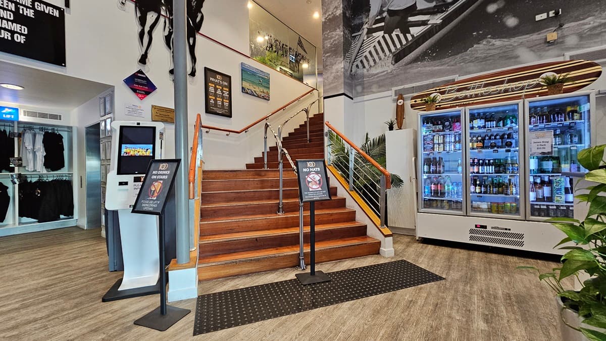 On the left, there is a kiosk with a touchscreen and a sign above it, next to a display of black clothing. In the middle, a wooden staircase with a metal handrail leads to an upper level, with surf-themed artwork and a large black-and-white mural of surfers on the wall above. On the right, there is a large refrigerated display case filled with drinks, and a potted plant beside it. The floor is wooden with a black mat at the base of the stairs, and the ceiling has recessed lighting.