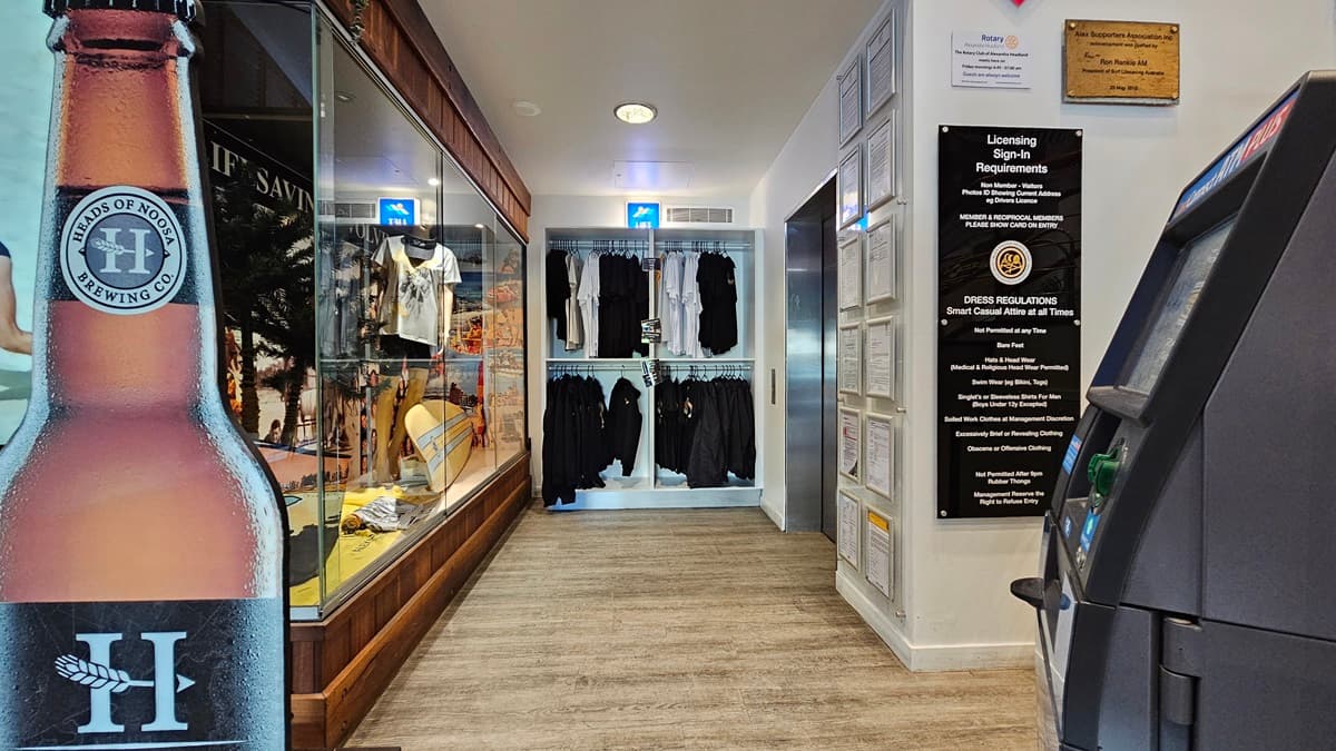On the left, there is a large poster of a beer bottle next to a glass display case containing various items, including clothing and surfboards. In the middle, there are shelves with neatly arranged black clothing. On the right, there is an ATM machine and a wall with licensing signs and requirements. The floor is wooden, and the ceiling has recessed lighting and a smoke detector.