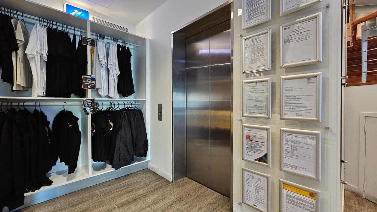 On the left, there are shelves with neatly arranged black and white clothing on hangers. In the middle, there is a stainless steel lift with closed doors and a control panel on the wall beside it. On the right, there is a wall with multiple framed documents and certificates. The floor is wooden, and the ceiling has recessed lighting and an air vent.