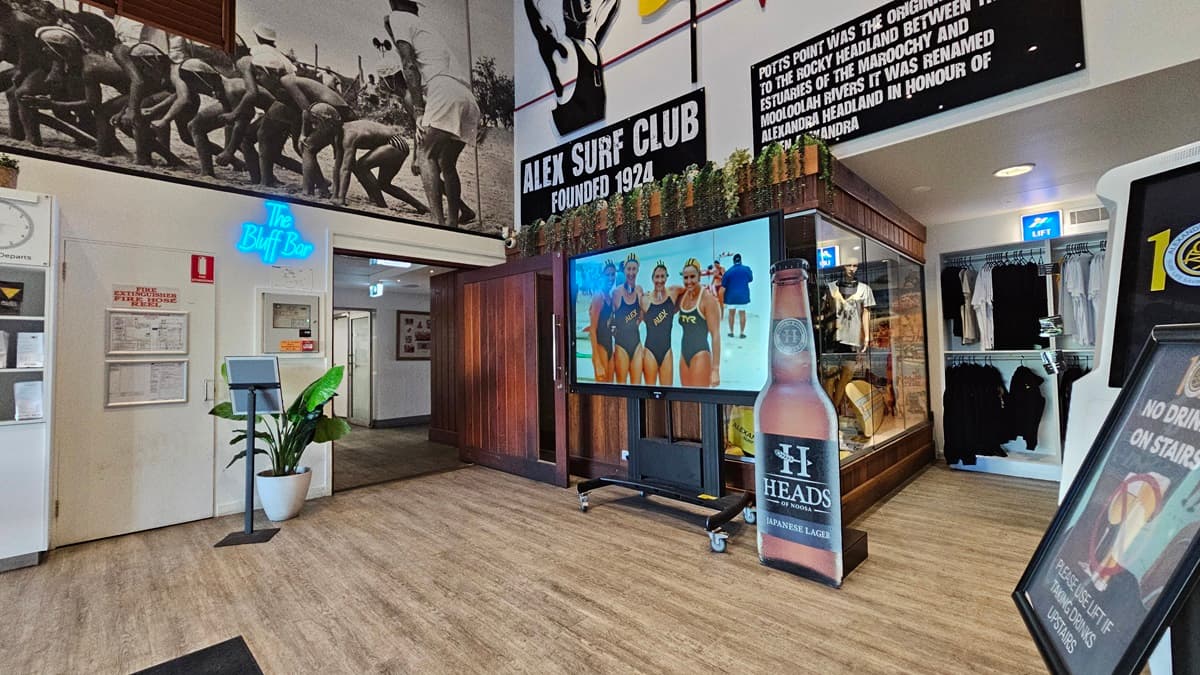 On the left, there is a white wall with a blue neon sign reading "The Buff Bar" above a small counter with a plant beside it. In the middle, there is a large screen on a stand displaying an image of people in swimsuits, with a surf club mural above it. On the right, there is a glass display case with surf club memorabilia and a blackboard with writing. The floor is wooden, and the ceiling has recessed lighting and surf club flags.