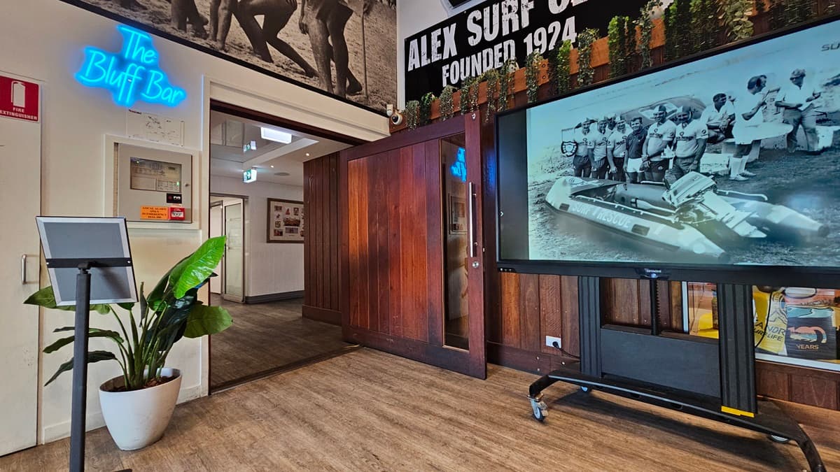 On the left, there is a white wall with a blue neon sign reading "The Bluff Bar" above a small counter with a plant beside it. In the middle, there is a wooden doorway leading to a hallway with framed photos on the walls. On the right, there is a large screen on a stand displaying a black-and-white image of people with a boat, and a glass display case with surf club memorabilia below it. The floor is wooden, and the ceiling has recessed lighting and surf club flags.