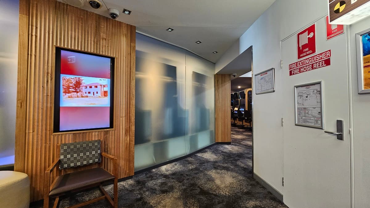 On the left, there is a wooden wall with vertical slats and a digital screen displaying an image of a building. Below the screen, there is a wooden chair with a patterned cushion. On the right, there is a white wall with fire safety signs, a fire extinguisher, and a door. In the middle, there is a frosted glass partition leading to a hallway with more chairs. The floor is carpeted with a dark, mottled pattern, and the ceiling has white panels with recessed lighting.