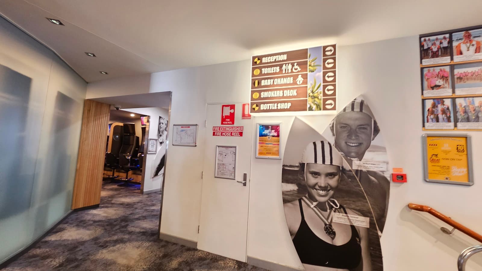 On the left, there is a wall with black and white photos of people and a wooden floor leading to a hallway. On the right, there is an ATM machine with a small trash bin next to it. In the middle, there is a decorative partition with circular cutouts. The floor is a mix of wooden and dark carpeted areas, and the ceiling has recessed lighting.