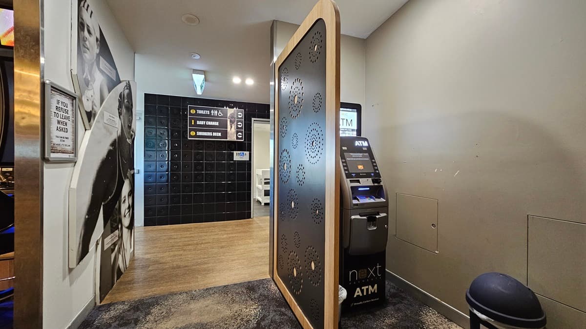 On the left, there is a wall with black and white photos of people and a wooden floor leading to a hallway. On the right, there is an ATM machine with a small trash bin next to it. In the middle, there is a decorative partition with circular cutouts. The floor is a mix of wooden and dark carpeted areas, and the ceiling has recessed lighting.