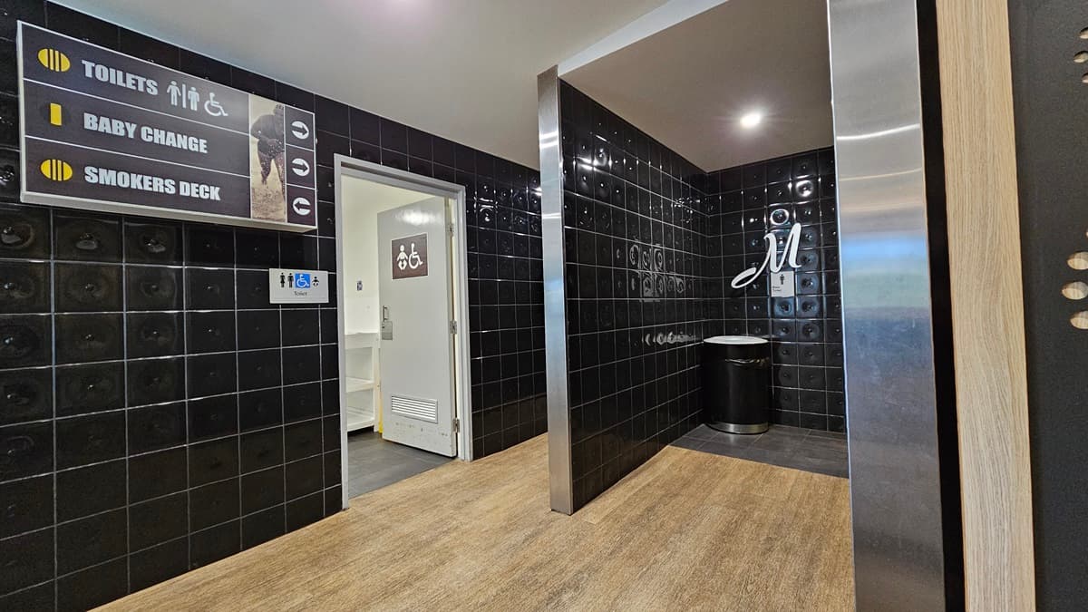 On the left, there is a sign indicating toilets, baby change, and smokers deck, above a doorway leading to a baby change room. On the right, there is a black tiled wall with a silver bin and a sign with the letter "M" for men's toilets. In the middle, there is a corridor with black tiled walls and a wooden floor. The ceiling is grey with recessed lighting.