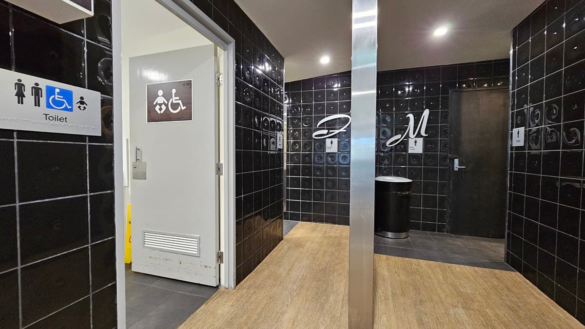 On the left, there is a doorway with a sign indicating accessible toilets, baby change, and a smokers' deck, leading to a baby change room with a white door. On the right, there is a black tiled wall with a silver bin and a sign with the letter "M" for men's toilets next to a black door. In the middle, there is a corridor with black tiled walls and a wooden floor. The ceiling is grey with recessed lighting.