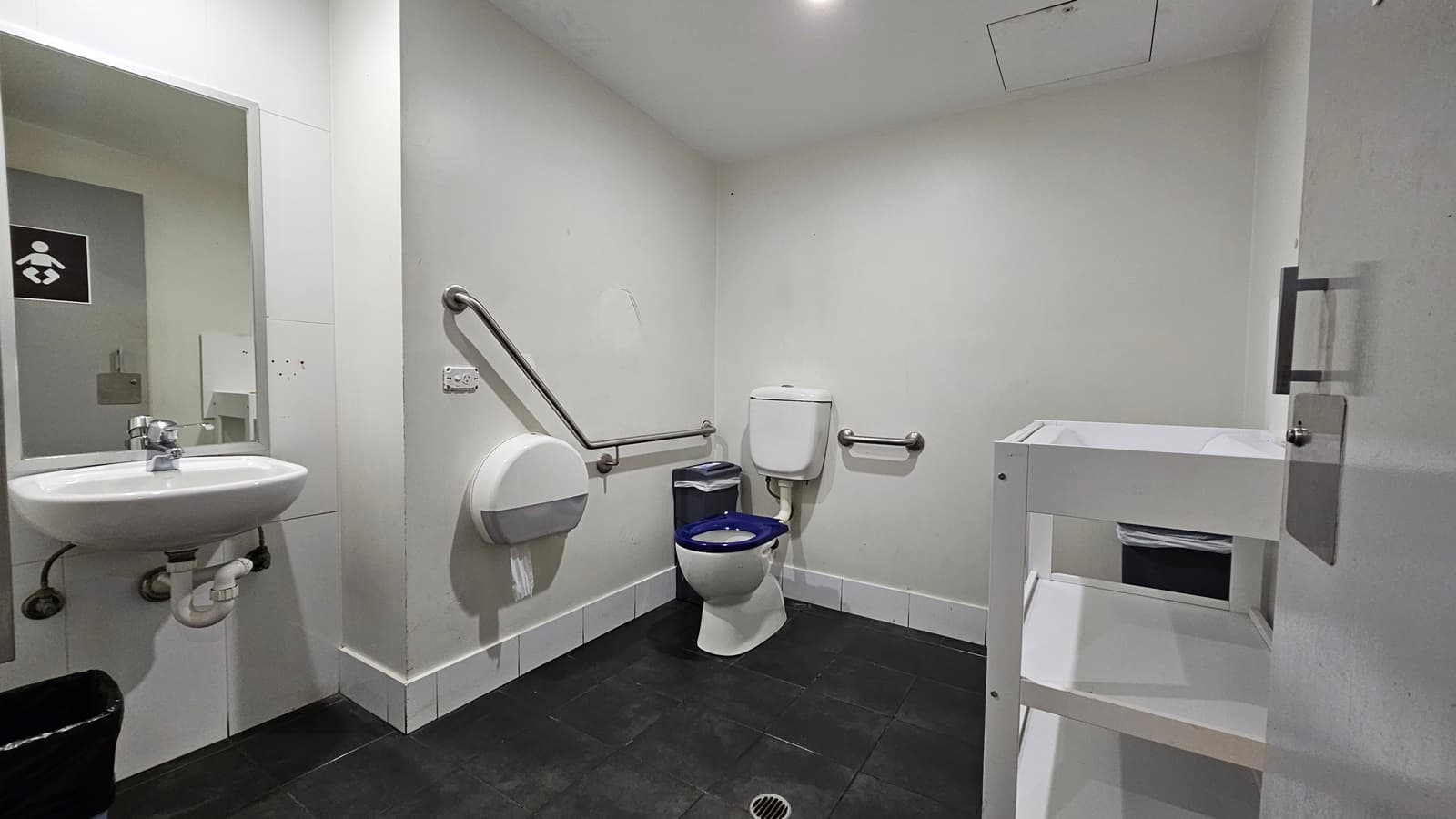 On the left, there is a white sink with a mirror above it and a black bin below. Next to the sink, there is a wall-mounted baby changing station. On the right, there is a white shelving unit with a bin on the bottom shelf. In the middle, there is a toilet with a blue seat, a grab rail on the wall, and a toilet paper dispenser. The floor is covered with black tiles, and the ceiling is white with recessed lighting.