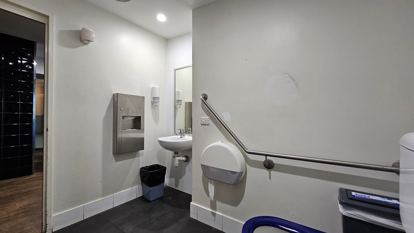On the left, there is a doorway leading to a hallway with dark tiles. Next to the doorway, there is a wall-mounted paper towel dispenser above a black bin. Further right, there is a white sink with a mirror above it and soap dispensers on either side. On the right, there is a grab rail mounted on the wall, a wall-mounted baby changing station, and a toilet with a blue seat. The floor is covered with dark tiles, and the ceiling is white with recessed lighting.