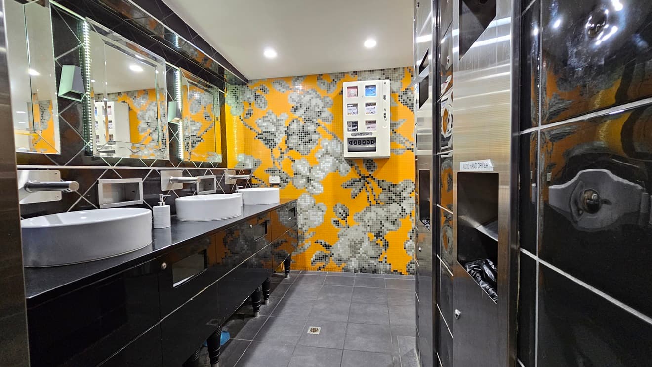 On the left, there are two white sinks with mirrors above them, set on a black countertop with soap dispensers. Below the countertop, there are storage shelves. On the right, there is a black tiled wall with a silver hand dryer and a vending machine. In the background, the wall is covered with orange and grey floral mosaic tiles. The floor is covered with dark grey tiles, and the ceiling is white with recessed lighting.
