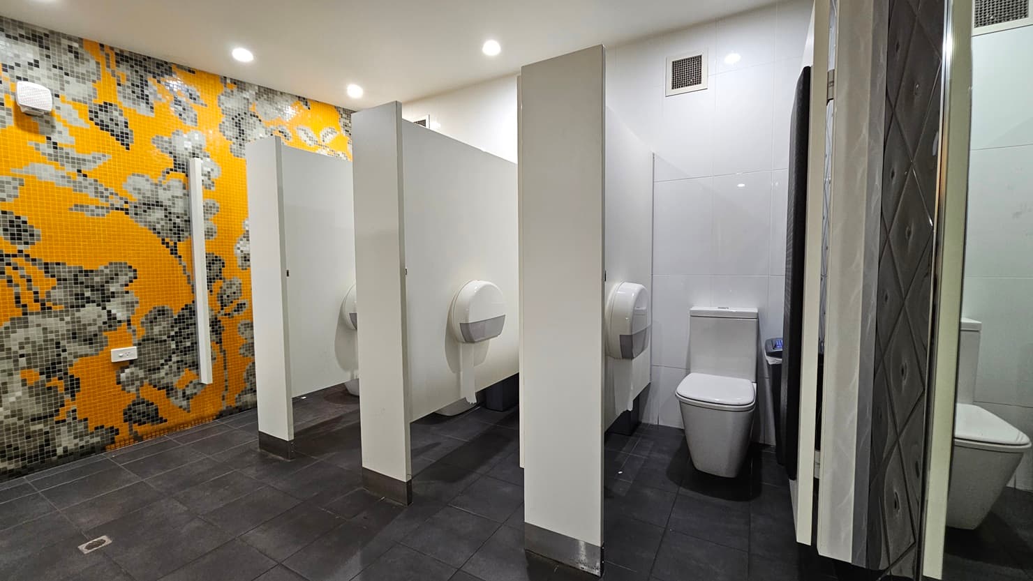 On the left, there is a wall covered with orange and grey floral mosaic tiles. In the middle, there are three white toilet cubicles with partial walls and doors, each containing a toilet and a large toilet paper dispenser. On the right, there is a white tiled wall with a full-length mirror. The floor is covered with dark grey tiles, and the ceiling is white with recessed lighting.