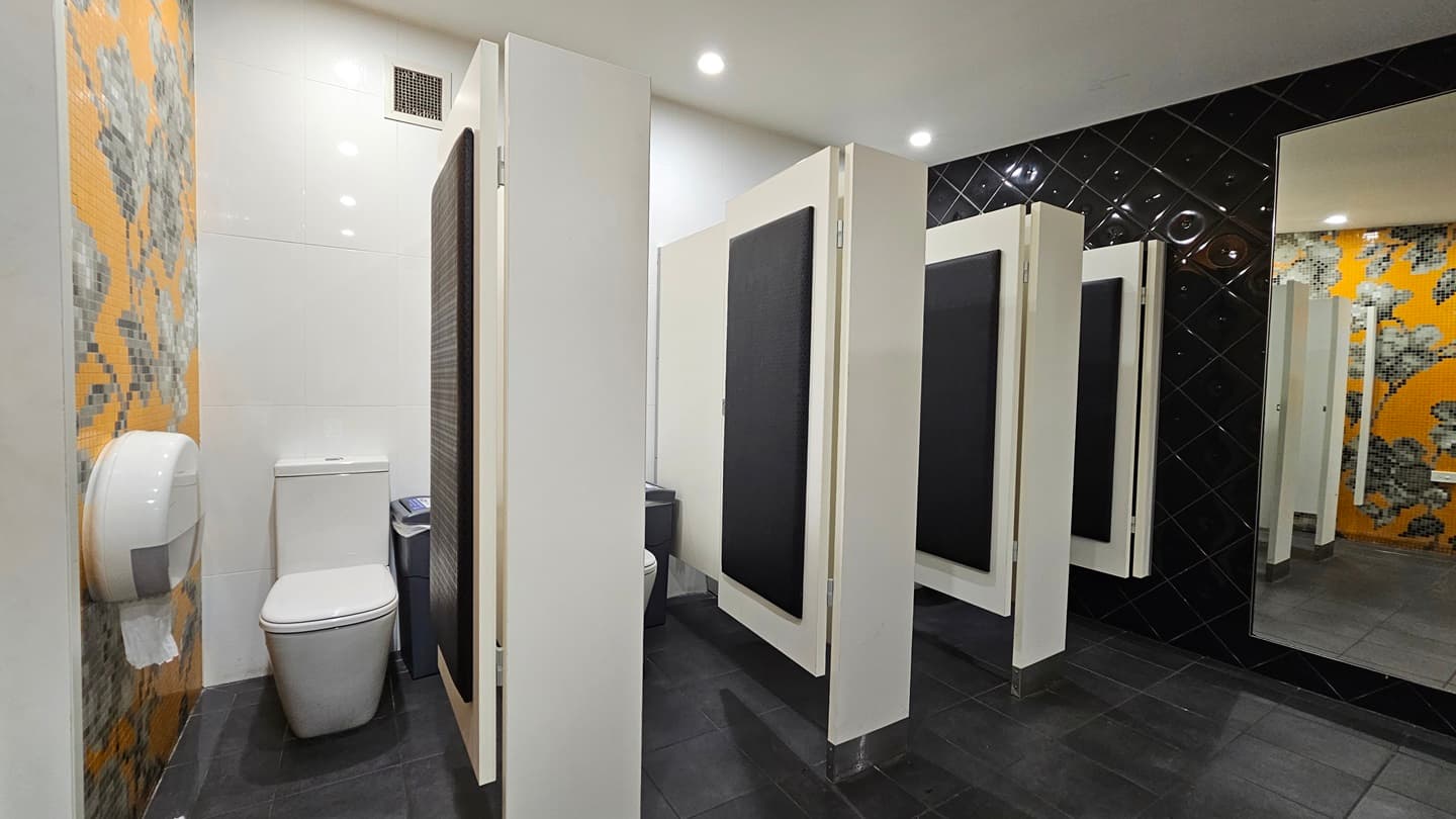 On the left, there is a toilet with a large toilet paper dispenser mounted on a wall covered with orange and grey floral mosaic tiles. In the middle, there are three white toilet cubicles with black doors, each containing a toilet. On the right, there is a black tiled wall with a full-length mirror. The floor is covered with dark grey tiles, and the ceiling is white with recessed lighting.