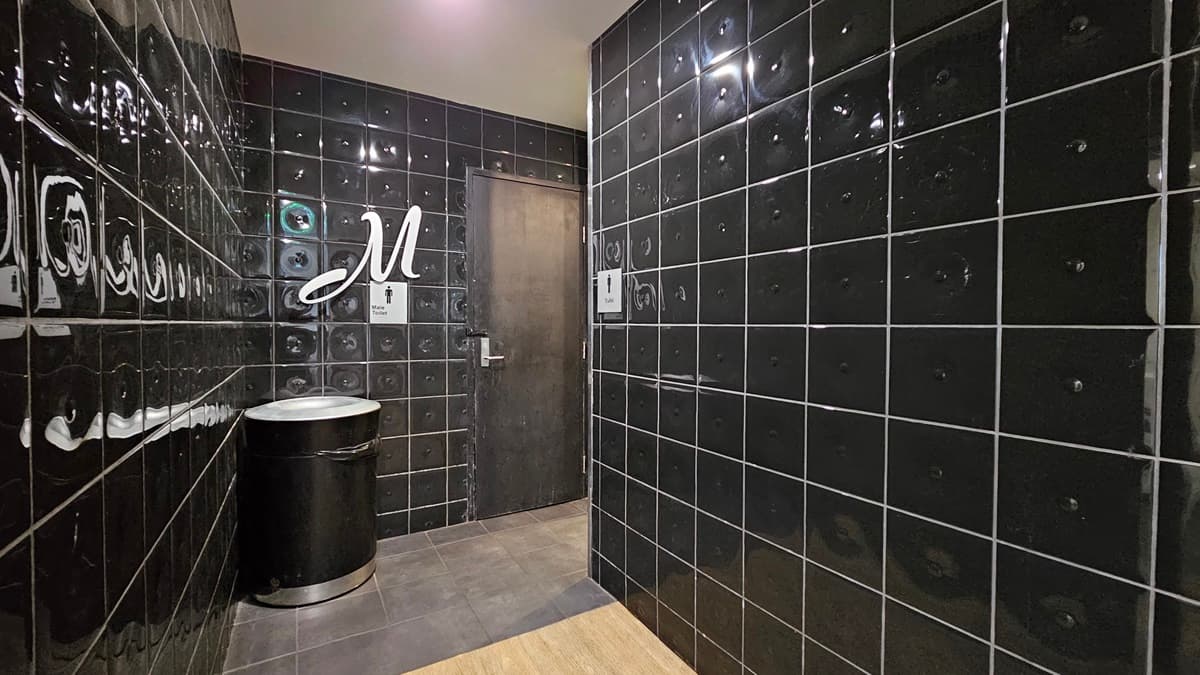 On the left, there is a black tiled wall with a silver bin and a sign with the letter "M" above a door. In the middle, there is a dark wooden door with a handle. On the right, there is another black tiled wall. The floor is covered with dark grey tiles, and the ceiling is white with recessed lighting.