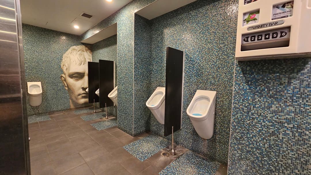 On the left, there is a wall with a large artistic face mural. In the middle, there are three white urinals separated by black dividers, mounted on a wall covered with blue mosaic tiles. On the right, there is a vending machine for toiletries attached to the same blue mosaic-tiled wall. The floor is covered with dark grey tiles, and the ceiling is white with recessed lighting.