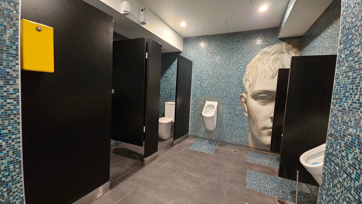 On the left, there are black cubicle doors with a yellow panel on one of them. In the middle, there is a white urinal mounted on a blue mosaic-tiled wall featuring a large mural of a face. On the right, there is a white sink with a blue mosaic-tiled backsplash. The floor is covered with dark grey tiles, and the ceiling is white with recessed lighting.