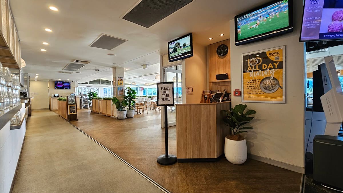 On the left, there is a long counter with a display case and a row of overhead lights. In the middle, there is a wooden reception desk with a sign that reads "Please Wait to be Seated," and a potted plant beside it. In the background, there are dining tables with chairs and another counter area. On the right, there are two wall-mounted screens, one showing a sports channel and the other displaying a menu. The floor is a mix of carpet and wood, and the ceiling has recessed lighting.