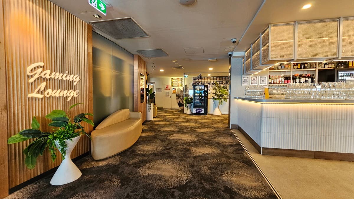On the left, there is a wooden wall with a neon sign reading "Gaming Lounge" and a white sofa next to potted plants. In the middle, there is a hallway with a dark, patterned carpet leading to a vending machine and a door in the background. On the right, there is a long bar counter with stools, shelves stocked with bottles, and glasses hanging overhead. The ceiling has recessed lighting and air conditioning vents.