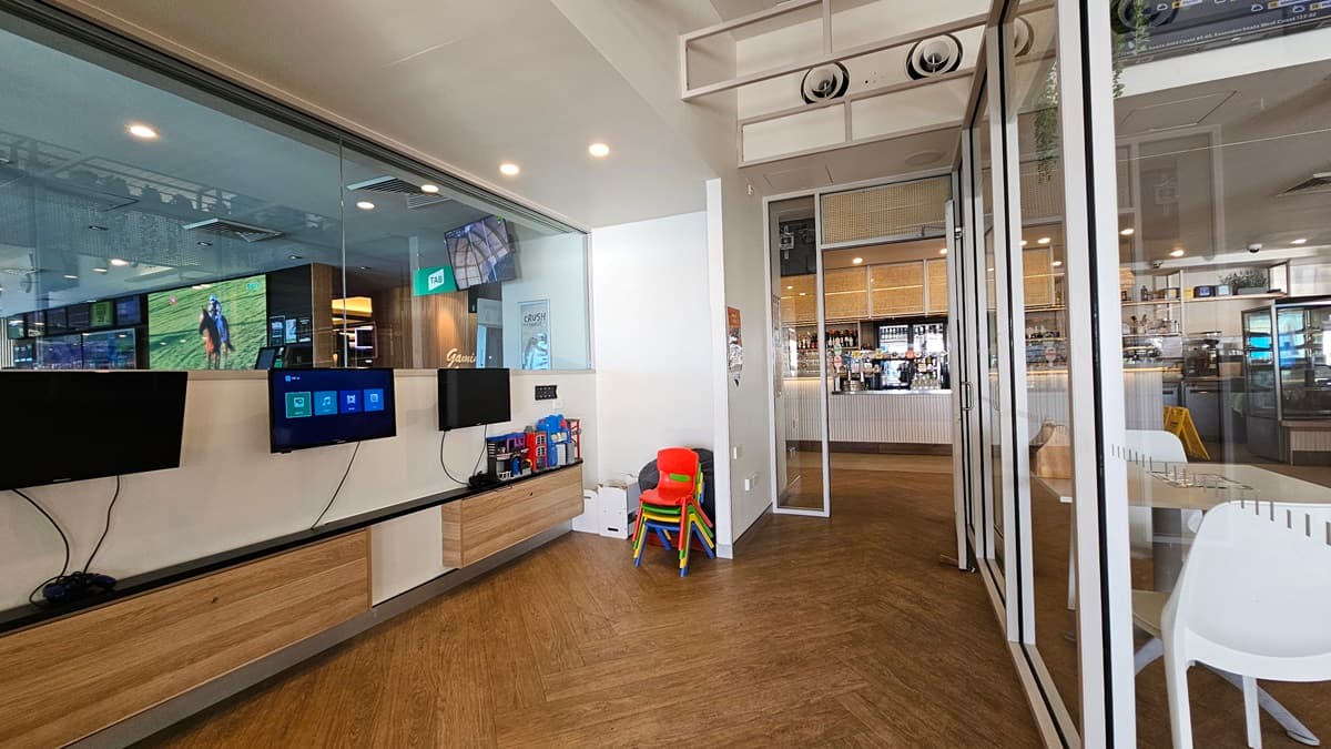 On the left, there are wall-mounted screens displaying various images and information, with wooden cabinets below them. In the middle, there is a small stack of colourful children's chairs and a red high chair. In the background, there is a bar area with stools, shelves stocked with bottles, and a counter. On the right, there are glass doors and windows leading to the bar area, with a white chair nearby. The floor is wooden, and the ceiling has recessed lighting and air conditioning vents.