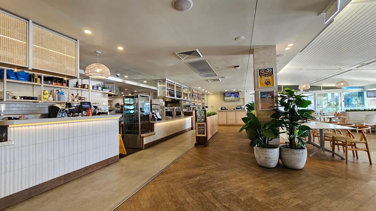 On the left, there is a long counter with a white front, illuminated from below, and a display case with pastries. Behind the counter are shelves with bottles and coffee machines. In the middle, there is a wooden partition with potted plants in front of it. In the background, there is another counter area with a digital screen above it. On the right, there are tables and chairs near large windows. The floor is wooden, and the ceiling has recessed lighting and air conditioning vents.