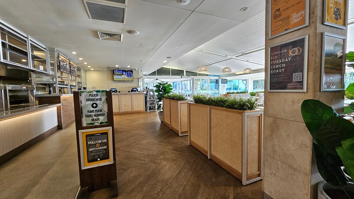 On the left, there is a long counter with a white front, a "Please Order Here" sign, and shelves with various items above. In the middle, there is a wooden partition with potted plants on top, leading to a dining area with tables and chairs near large windows. In the background, there is a digital screen and more seating. On the right, there are posters on the wall, a potted plant, and a sign about a lunch roast. The floor is wooden, and the ceiling has recessed lighting and air con vents.