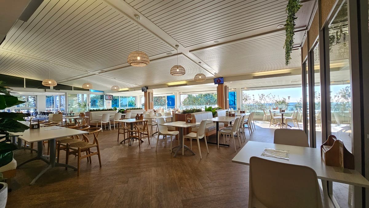 On the left, there are several tables and chairs near large windows with a view of the sea. In the middle, there are more tables and chairs, with a wooden partition featuring potted plants on top. In the background, there are additional seating areas and large windows showcasing the sea view. On the right, there are more tables and chairs near floor-to-ceiling windows. The floor is wooden, and the ceiling has white panels with recessed lighting and hanging wicker lamps.