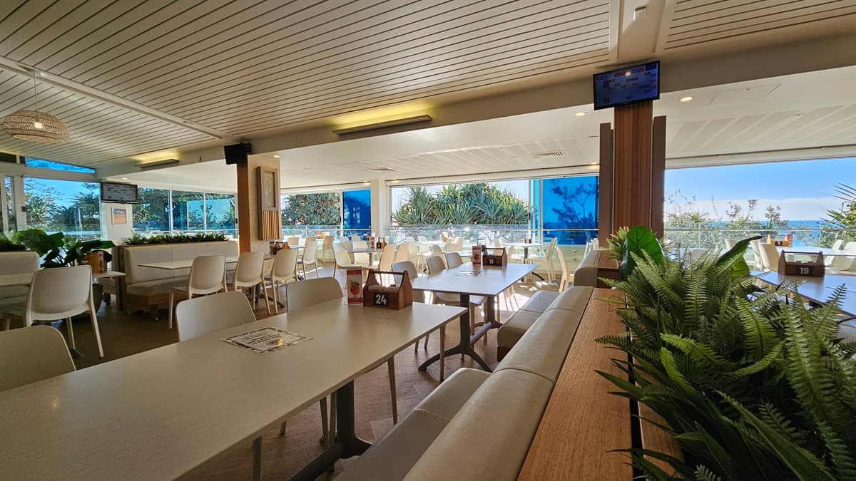 On the left, there are white tables and chairs near large windows with a view of trees and foliage. In the middle, there are more tables and chairs, with a wooden partition featuring potted plants on top. In the background, there are additional seating areas and large windows with blue blinds. On the right, there are more tables and chairs near floor-to-ceiling windows. The floor is wooden, and the ceiling has white panels with recessed lighting and a mounted TV screen.