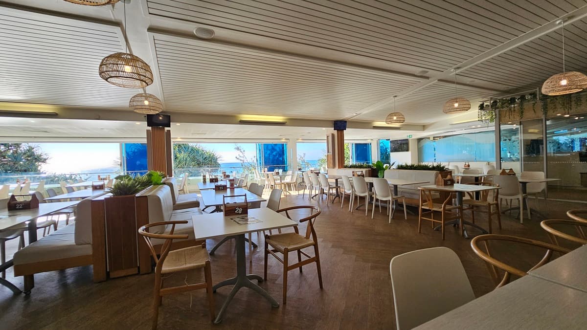 On the left, there are white tables and chairs near large windows with a view of the sea and trees. In the middle, there are more tables and chairs, with a wooden partition, potted plants on top. In the background, there are additional seating areas and large windows with blue blinds. On the right, there are more tables and chairs near floor-to-ceiling windows, with a section of the room separated by glass walls. The floor is wooden, and the ceiling has white panels with recessed lighting.