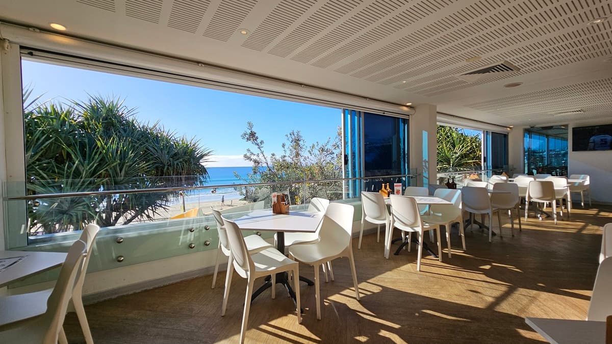 On the left, there are white tables and chairs near large windows with a view of the beach and trees. On the right, there are more white tables and chairs near floor-to-ceiling windows, with blue blinds partially drawn. In the middle, there are additional tables and chairs, with sunlight streaming through the windows. The floor is wooden, and the ceiling has white panels with recessed lighting and air conditioning vents.