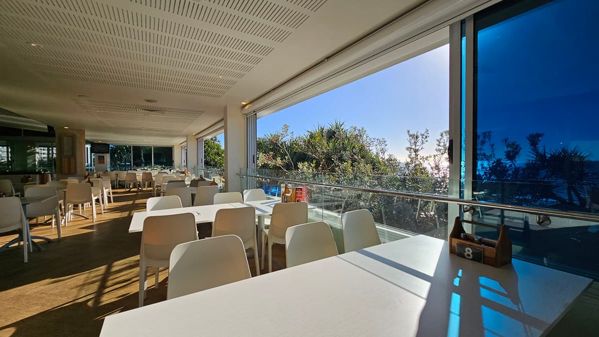 On the left, there are white tables and chairs near large windows with a view of the beach and trees. On the right, there are more white tables and chairs near floor-to-ceiling windows with blue-tinted glass. In the middle, there are additional tables and chairs, with a wooden partition featuring potted plants on top. The floor is wooden, and the ceiling has white panels with recessed lighting, air conditioning vents, and hanging wicker lamps.
