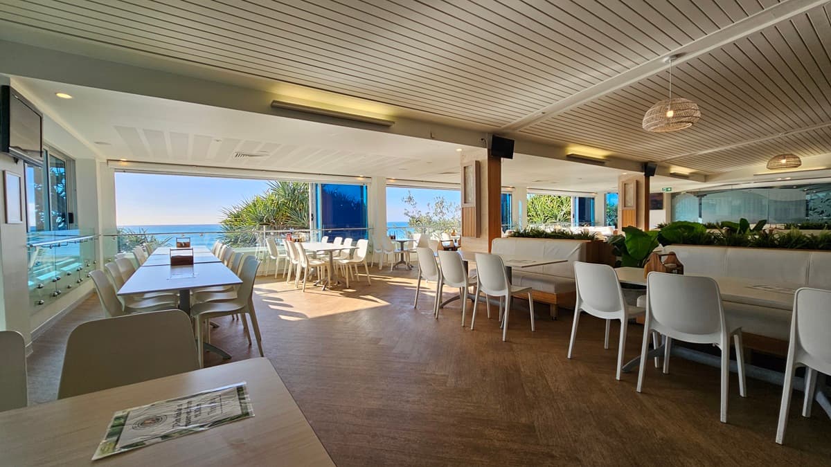 On the left, there are white tables and chairs near large windows with a view of the beach and trees. On the right, there are more white tables and chairs near floor-to-ceiling windows with blue-tinted glass. In the foreground, there are additional tables and chairs, with a wooden partition featuring potted plants on top. The floor is wooden, and the ceiling has white panels with recessed lighting, air conditioning vents, and hanging wicker lamps.
