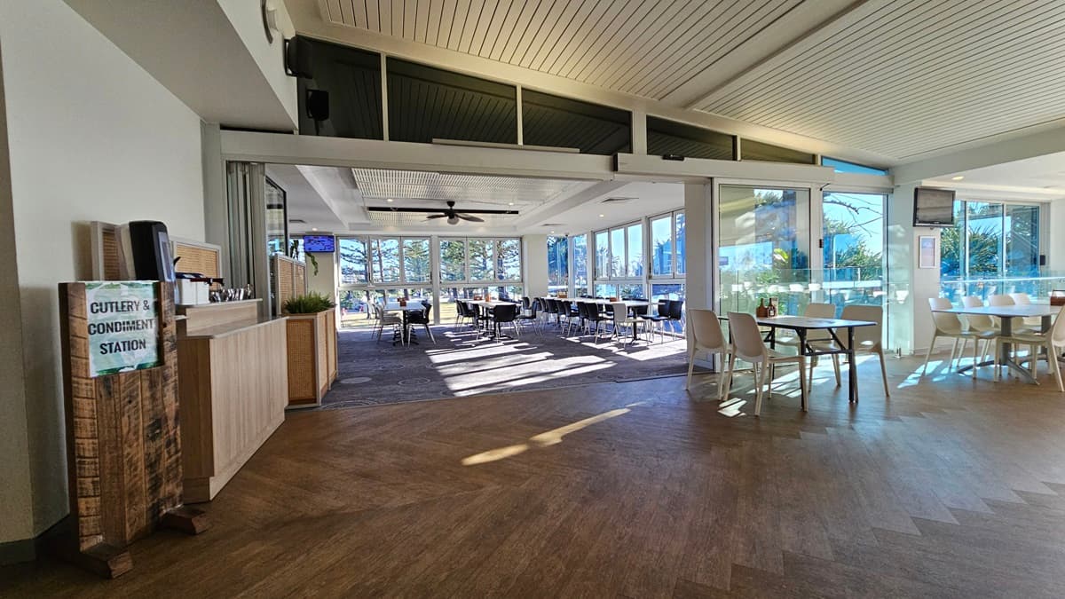 On the left, there is a wooden cutlery and condiment station next to a light wood counter. On the right, there are white tables and chairs near floor-to-ceiling windows with a view of trees. In the background, there are more tables and chairs, with large windows providing additional views of trees. The floor is a mix of wooden and carpeted areas, and the ceiling has white panels with recessed lighting and a ceiling fan.