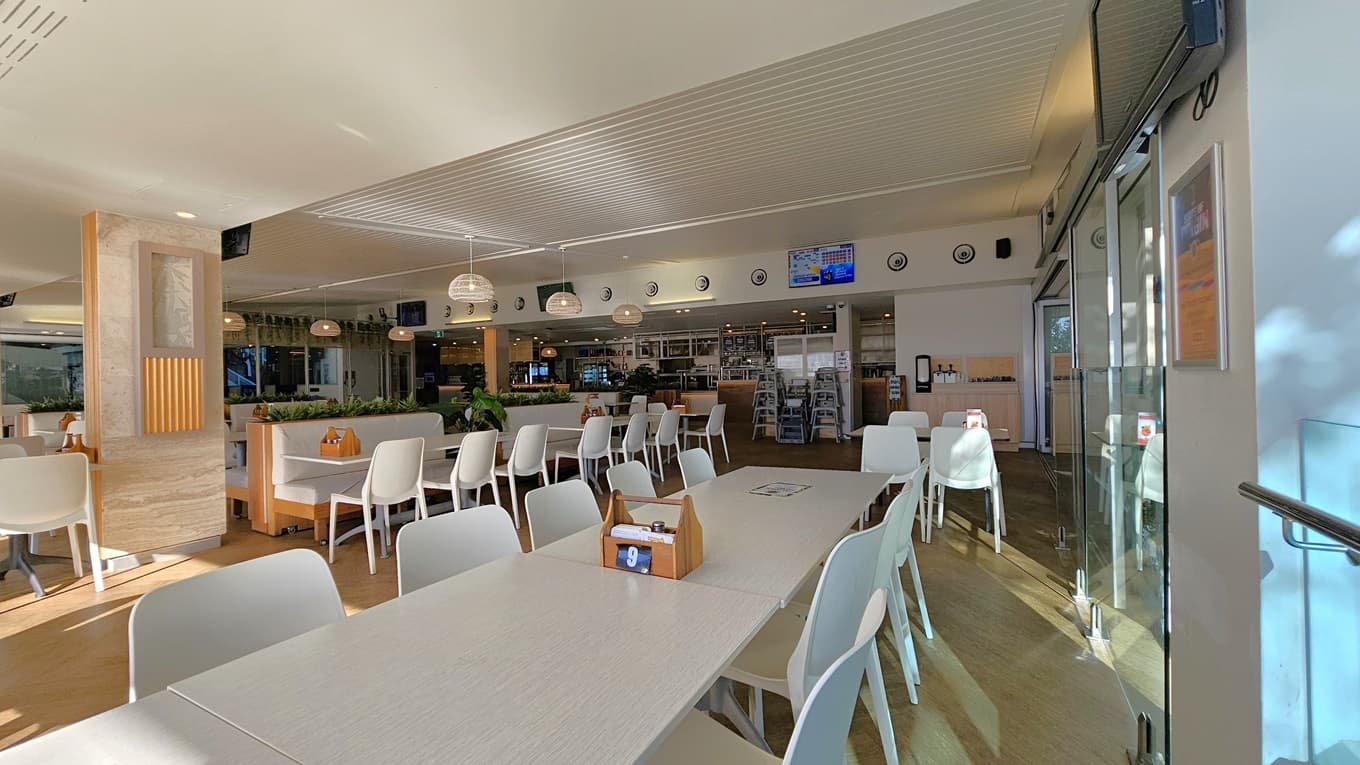 On the left, there are white tables and chairs near a wooden partition with potted plants on top. On the right, there are more white tables and chairs near floor-to-ceiling windows with a view of trees. In the background, there is a counter with bar stools, a menu board, and a display of drinks. The floor is wooden, and the ceiling has white panels with recessed lighting and hanging spherical lamps.