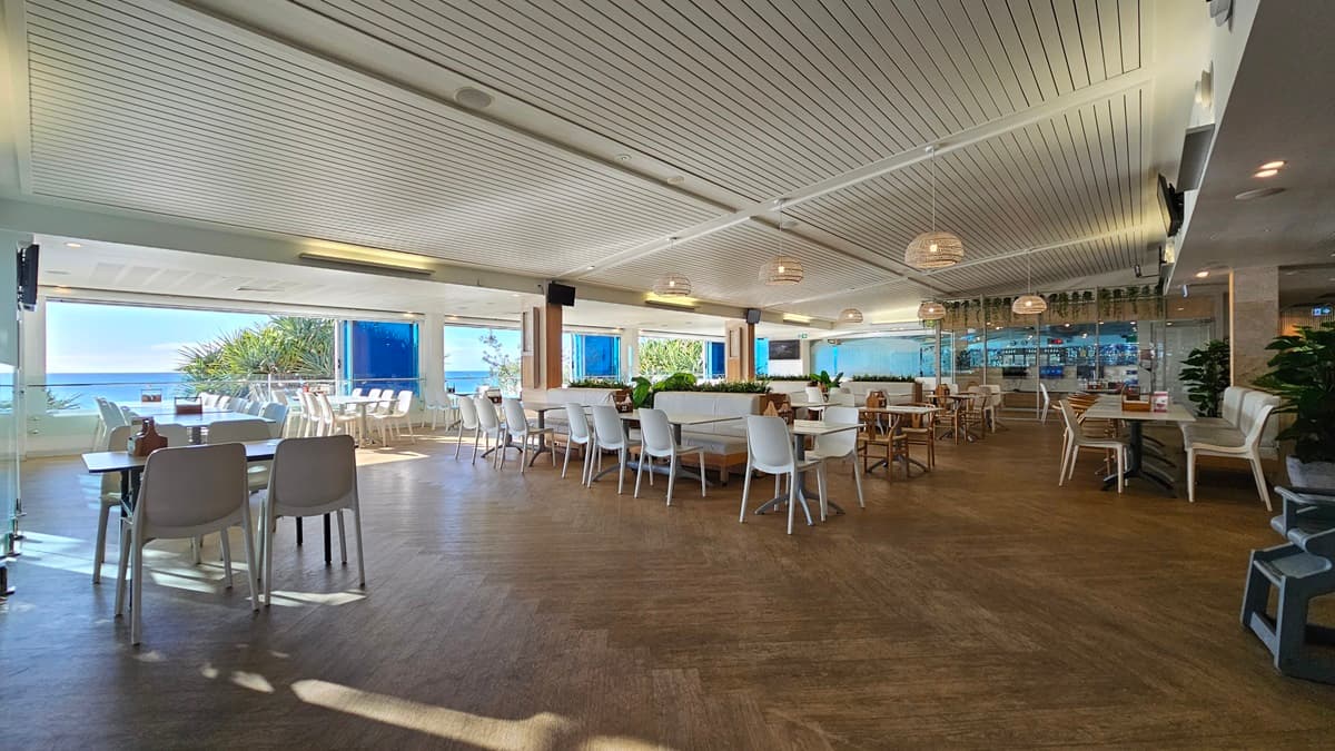 On the left, there are white tables and chairs near large windows with a view of the beach and trees. On the right, there is a bar area with stools, shelves of bottles, and more white tables and chairs. In the middle, there are additional tables and chairs, with a circular wooden partition featuring potted plants. The floor is wooden, and the ceiling has white panels with recessed lighting and hanging spherical lamps.