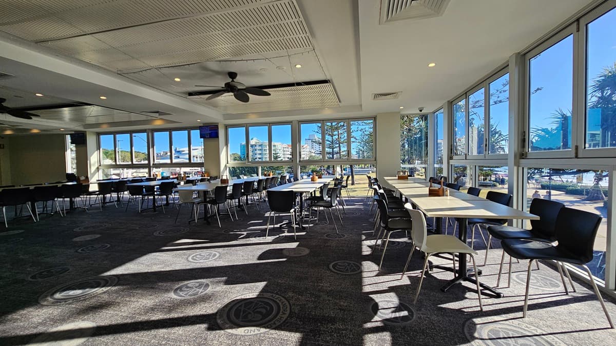 On the left, there are rows of black chairs and white tables near large windows. On the right, there are more white tables and black chairs near floor-to-ceiling windows with views of trees and buildings. In the middle, there are additional tables and chairs with sunlight streaming through the windows. The floor is carpeted with circular patterns, and the ceiling has white panels with recessed lighting and ceiling fans.