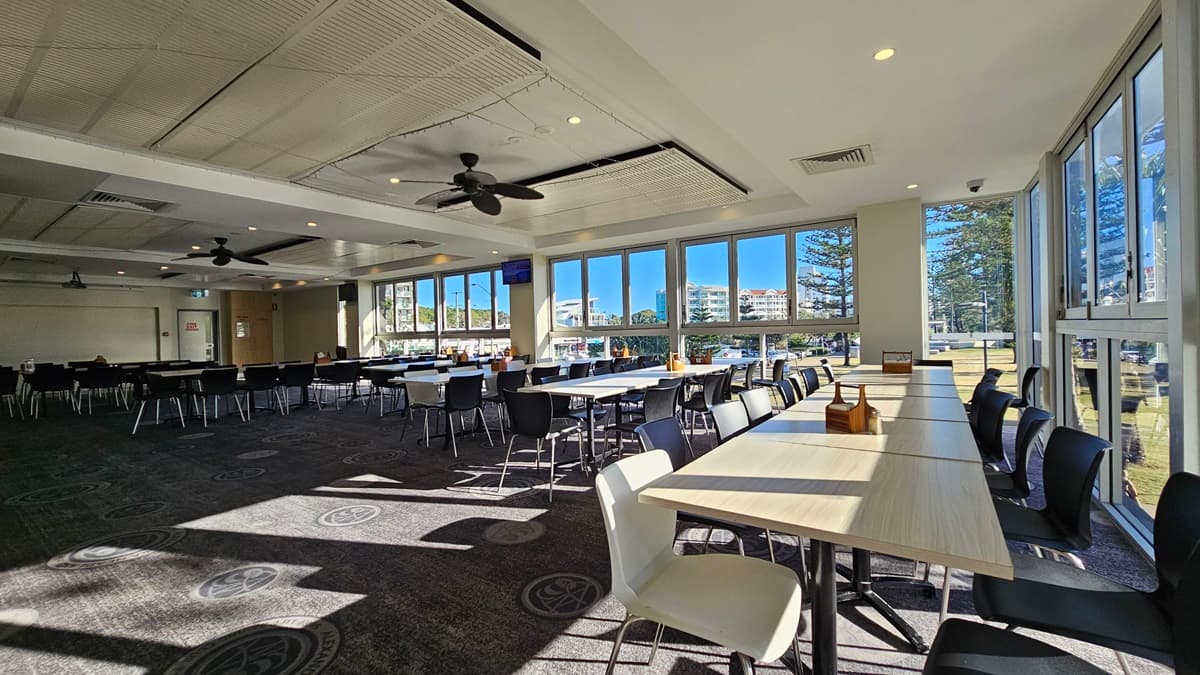 On the left, there are rows of black chairs and white tables near large windows. On the right, there are more white tables and black chairs near floor-to-ceiling windows with views of trees and buildings. In the middle, there are additional tables and chairs with sunlight streaming through the windows. The floor is carpeted with circular patterns, and the ceiling has white panels with recessed lighting and ceiling fans.