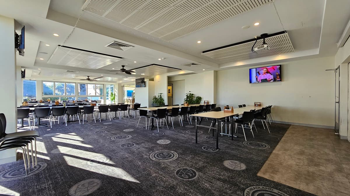 On the left, there are rows of black chairs and white tables near large windows with sunlight streaming in. On the right, there are more black chairs and white tables, with a television mounted on the wall displaying a programme. In the background, there is a bar area with stools and additional seating. The floor is carpeted with circular patterns, and the ceiling has white panels with recessed lighting, air conditioning vents, and ceiling fans.