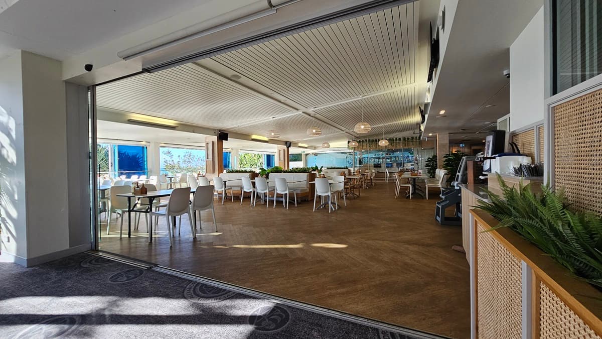 On the left, there are white tables and chairs near large windows with a view of trees and the sea. On the right, there is a wooden counter with potted plants and a coffee machine. In the middle, there are more white tables and chairs arranged in rows. The floor is a mix of wooden and carpeted areas, and the ceiling has white panels with recessed lighting and hanging spherical lamps.