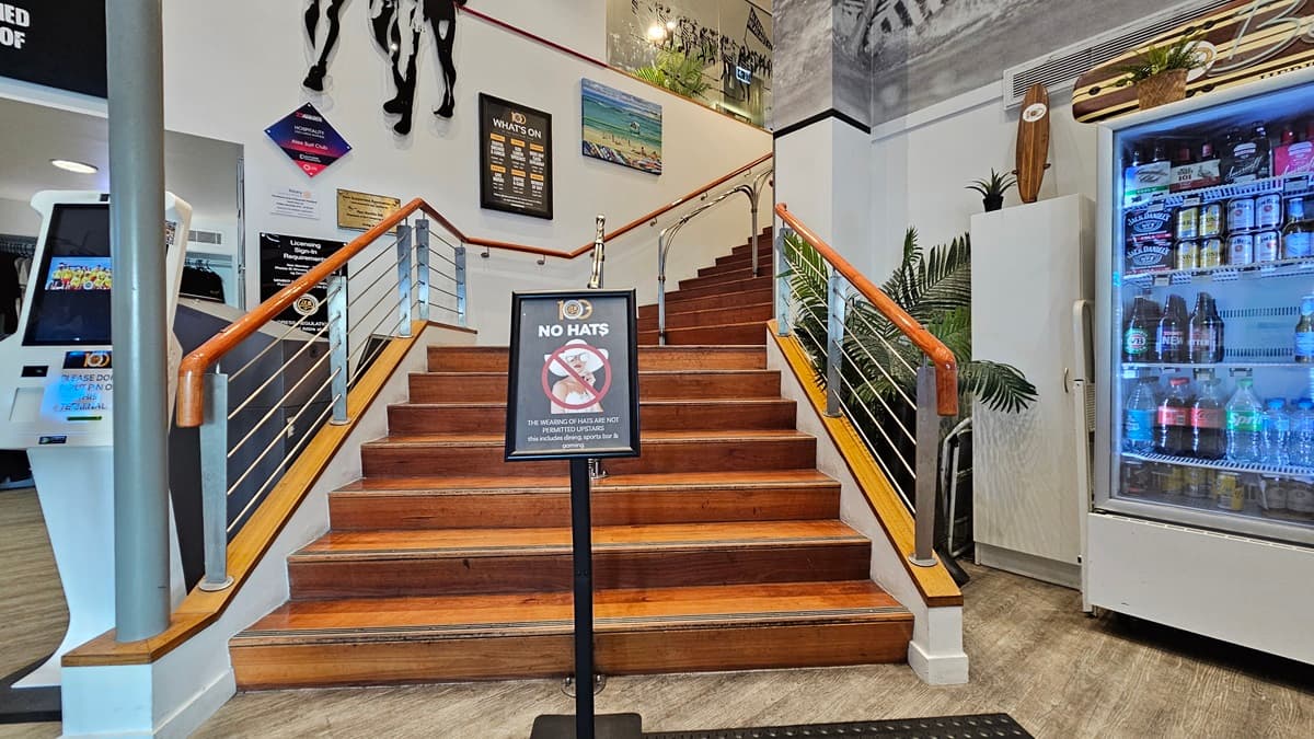 On the left, there is a white kiosk with a touchscreen and a wall with various signs. In the middle, there is a wooden staircase with metal railings leading up to a landing, with a "No Hats" sign at the base. In the background, there are framed pictures and a plant on the landing. On the right, there is a large refrigerator stocked with drinks and a tall potted plant beside it. The floor is light-coloured wood, and the ceiling has white panels with recessed lighting.