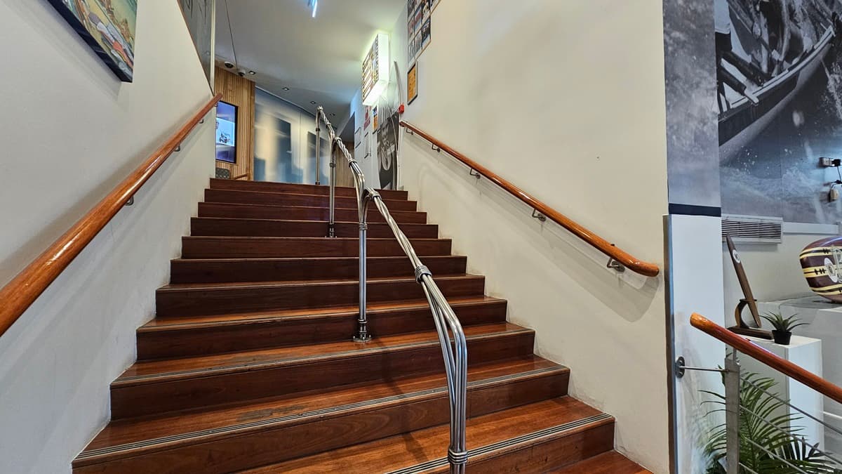On the left, there is a wooden handrail along the wall with framed pictures above it. In the middle, a wooden staircase with metal railings leads up to a landing, where a door and more framed pictures are visible. On the right, there is another wooden handrail, a large black-and-white mural on the wall, and a potted plant at the base of the stairs. The floor is wooden, and the ceiling has recessed lighting and a small light fixture near the top of the stairs.