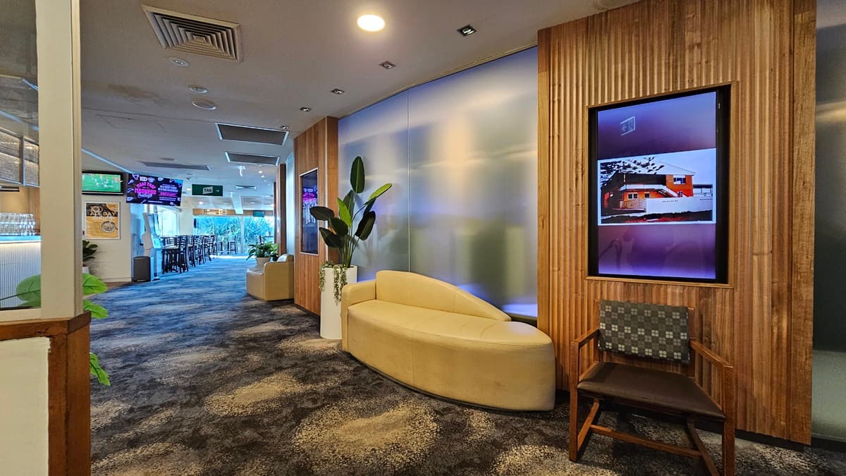 On the left, there is a hallway with a bar area visible in the distance, featuring bar stools and multiple wall-mounted TVs. In the middle, there is a frosted glass partition with a curved beige sofa and a tall potted plant in front of it. On the right, there is a wooden panelled wall with a framed digital display showing an image of a house, and a wooden chair with a patterned cushion. The floor is covered in dark, patterned carpet, and the ceiling has recessed lighting and air con vents.