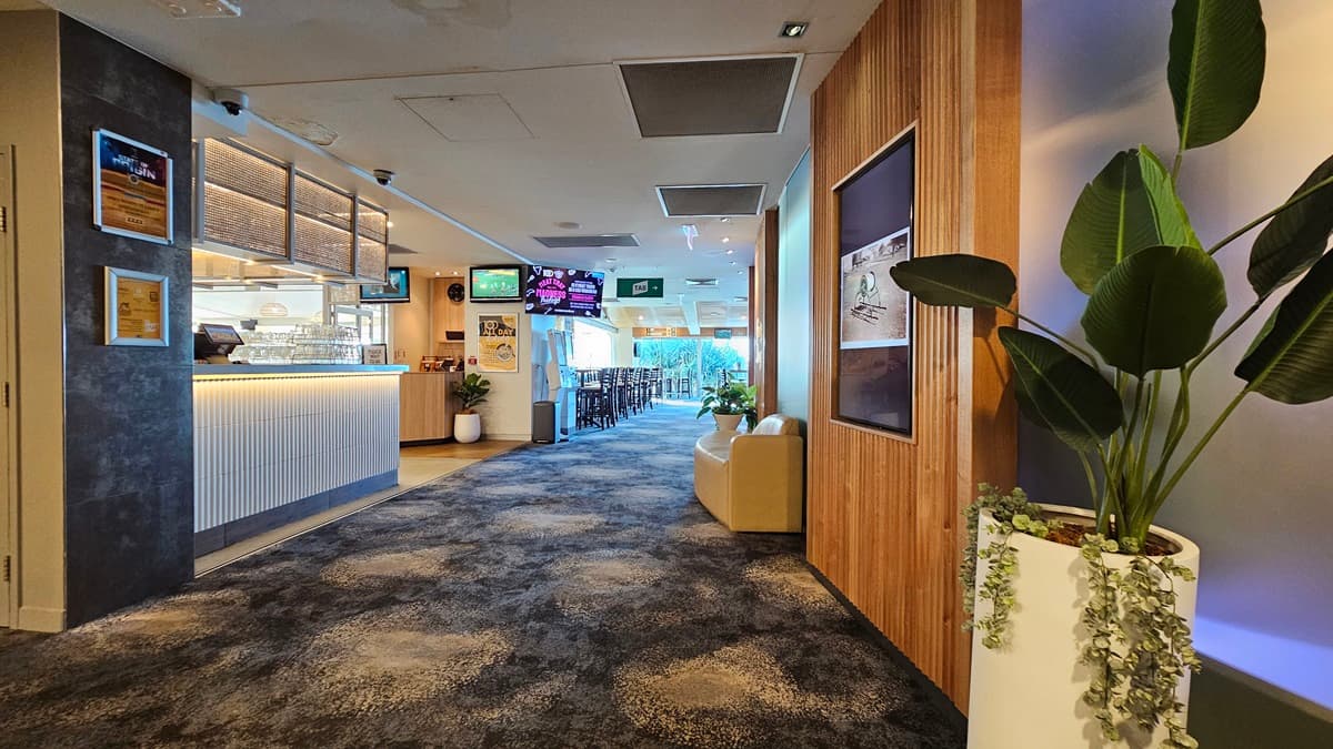 On the left, there is a bar area with a white counter, bar stools, and a menu board on the wall. In the middle, there is a hallway with a dark, patterned carpet leading to a seating area in the background, featuring tables and chairs. On the right, there is a wooden panelled wall with framed pictures, a beige sofa, and a tall potted plant. The ceiling has recessed lighting and air conditioning vents.